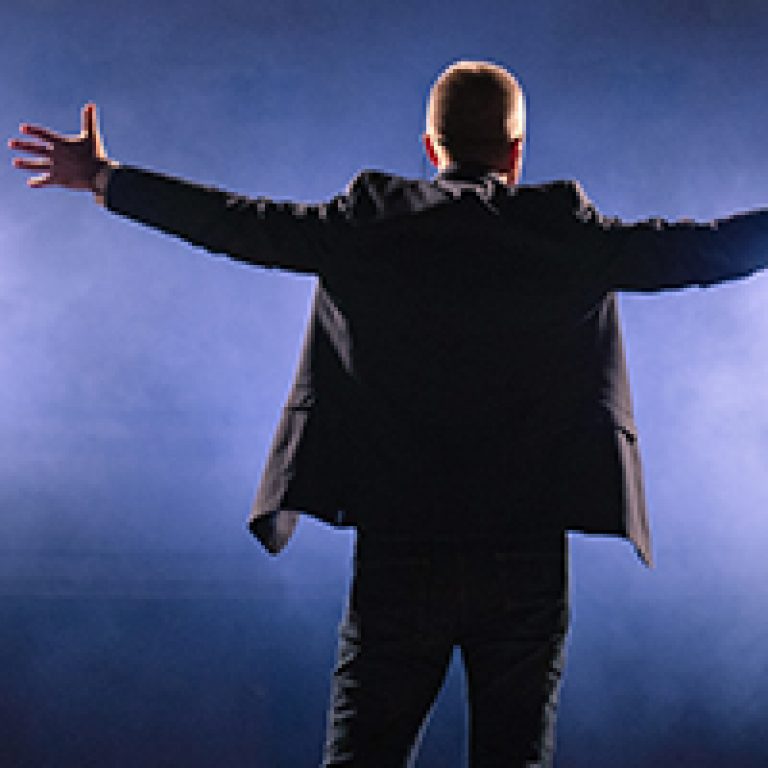 Showman. Middle Age Male entertainer, presenter or actor on stage. Arms to sides, smoke on background of spotlight. Rear view portrait of a male public speaker speaking at the microphone, pointing
